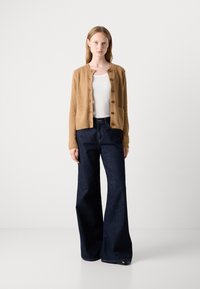 Cardigan beige con texture, colletto rotondo e chiusura con bottoni, abbinato a jeans ampi blu navy. Sotto una semplice maglietta bianca.