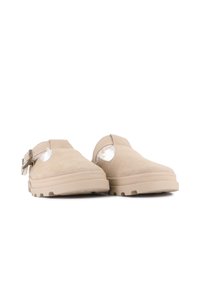 Beige mockasins i mocka med justerbara spännen och vit fluffig foder, mot en vit bakgrund.