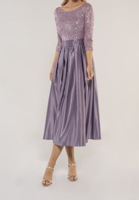 Ein fliederfarbenes Kleid mit einem besetzten Oberteil mit drei Viertelärmeln und einem plissierten Satinrock. Es hat eine taillierte Passform und wird mit metallischen High Heels kombiniert.