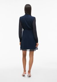 Γυναίκα που φοράει ένα γαλάζιο φόρεμα μέχρι το γόνατο με διαφανή μακριά μανίκια και λευκές ψηλοτάκουνες σάντες, στέκεται μπροστά σε λευκό φόντο.