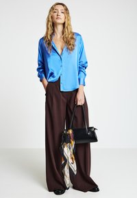 Woman standing wearing a shiny blue blouse, wide brown pants, holding a black handbag with a patterned scarf against a plain white background.