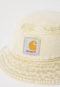 Gul bucket hat lavet af tekstureret stof, med et fremtrædende firkantet patch med et gult logo og "carhartt" tekst under.
