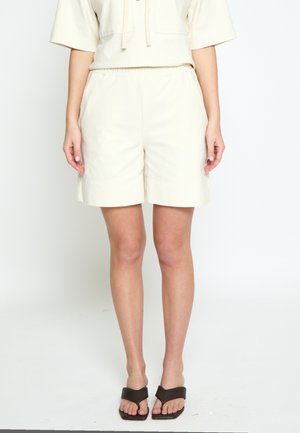 Naine kannab beežikat elastse taljega lühkseid pükse, sobiva lühikeste varrukatega särgi ja mustade ruudukujuliste varbavahedega sandaale, seistes ühevärvilise valge tausta ees.