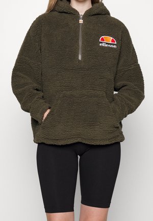 Osoba w ciemnozielonym, futrzanym bluzie z kapturem i suwakiem do ćwiartki oraz logo ellesse na piersi, w czarnych, rowerowych szortach.