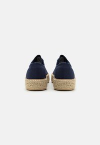 Navy canvas slip-on shoes with a textured, beige espadrille-style sole. Features subtle stitching and a rounded toe design.