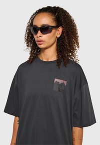 Czarny, oversize'owy t-shirt z małym graficznym wzorem na kieszeni; wykonany z miękkiego materiału, z krótkimi rękawami i luźnym krojem. Czarne okulary przeciwsłoneczne.