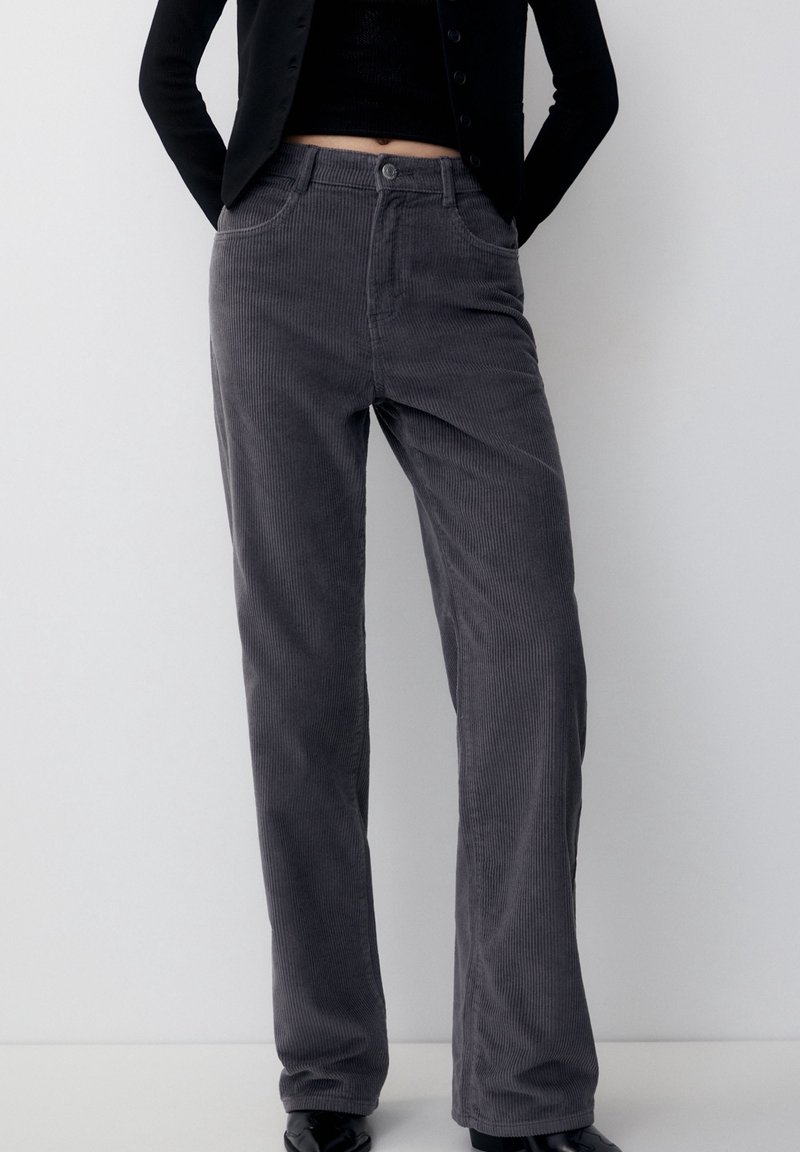 Personne portant un pantalon droit en velours côtelé gris foncé, un top court noir et une veste noire, debout devant un fond clair uni.
