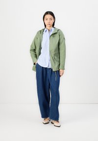 Olivengrønn hettejakke med snor, lyseblå stripete skjorte, vide blå denim-bukser og beige slip-on-sko.