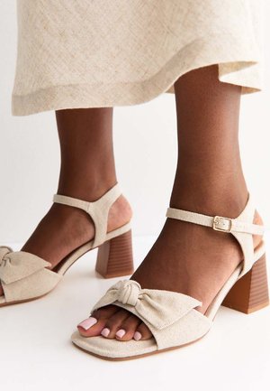 Pies de piel marrón que llevan sandalias beige de tacón ancho con tiras y detalle de lazo, combinadas con una falda ligera de tela.