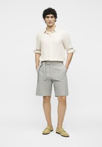 Homme portant une chemise beige à manches longues, un short gris avec ceinture et des mocassins jaunes, debout les mains dans les poches sur fond blanc.