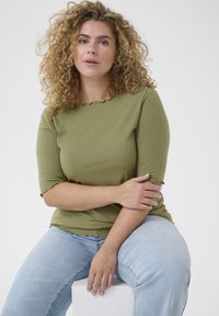 Haut côtelé vert olive avec un col en dentelle et des bords de manches scallopés, associé à un jean bleu clair. Modèle assis sur un bloc blanc.