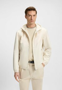 Giacca leggera con cappuccio e zip frontale, tasche laterali e pannelli a contrasto sulle spalle, indossata sopra un top beige e pantaloni.