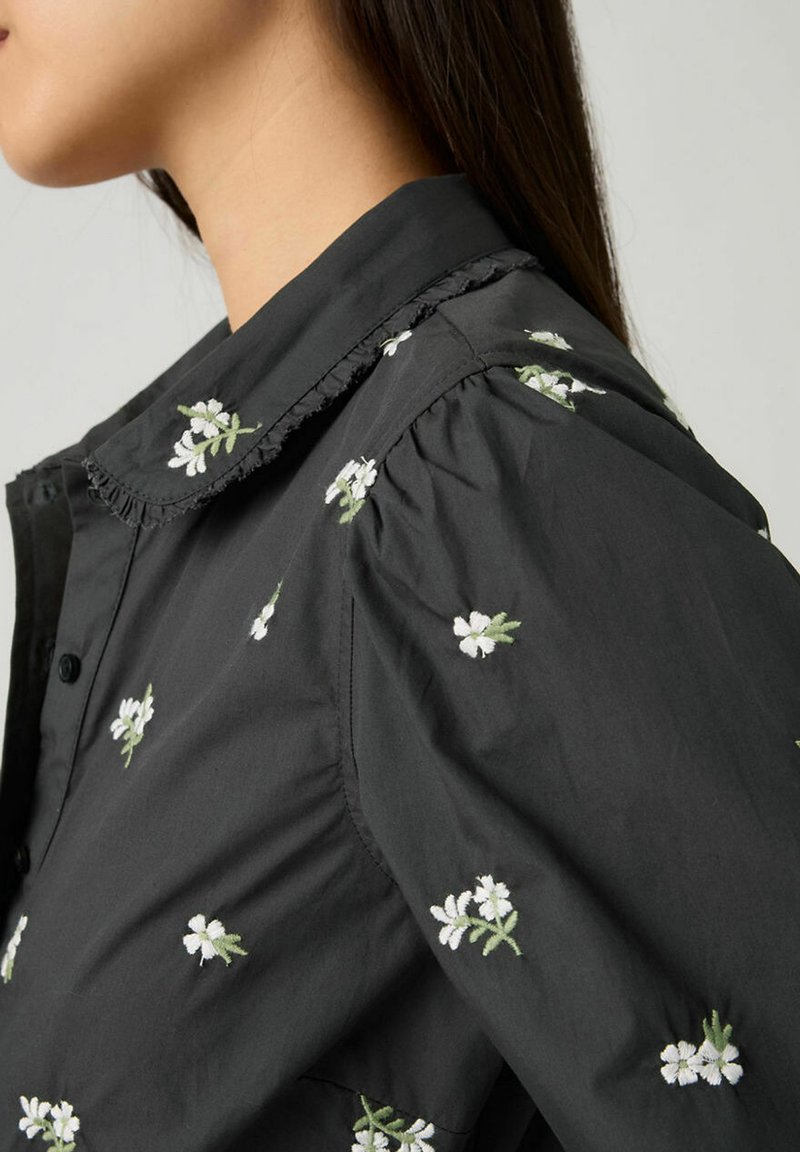 Blouse noire ornée de broderies florales blanches, avec un col à volants et des manches froncées, réalisée en tissu léger.