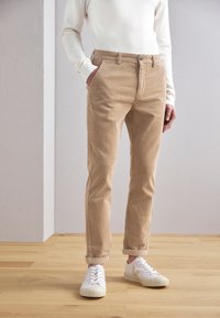Pantalon en velours côtelé beige, coupe slim, avec poches avant, fermeture par bouton, et ourlets roulés, associé à des baskets blanches.