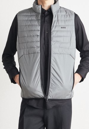 Mand iført en grå quiltet ærmeløs vest med BOSS-logo over en sort skjorte med knapper og sorte bukser, hænderne i vestens lommer.