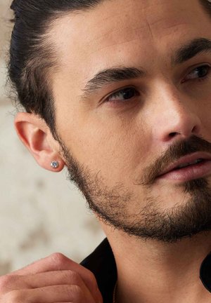 Silver stud earring featuring a clear gemstone, worn on a man’s ear. Close-up shows facial hair, skin texture, and a black shirt.