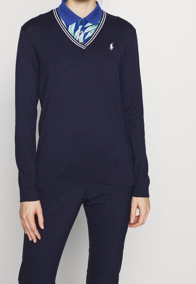 Personne portant un pull bleu marine en V par-dessus une chemise bleue à col avec un motif de feuilles vertes et blanches, associée à un pantalon bleu marine.