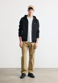 Jeune homme portant une casquette beige, un sweat à capuche zippé noir sur un t-shirt blanc, un pantalon beige et des baskets noires debout contre un mur blanc.