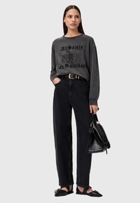 Donna in piedi che indossa una felpa stampata grigio scuro, pantaloni neri a vita alta, ballerine nere a punta e tiene in mano una borsa nera.