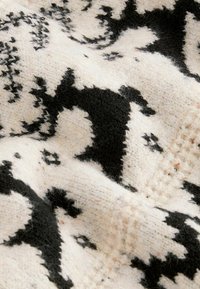 Wolldecke mit einem cremefarbenen Grund und schwarzen geometrischen Mustern. Die Textur wirkt weich, mit einem leicht flauschigen Finish und sichtbaren Naturfasern.