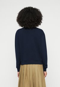 Tengerészkék pulóver kerek nyakkivágással, ejtett vállakkal és bordázott szegéllyel. Világos barna pliszírozott szoknya részben látható alul.