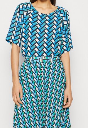 Femme portant un haut à manches courtes et un pantalon assortis avec un motif géométrique de maillons en bleu, vert, noir et blanc.