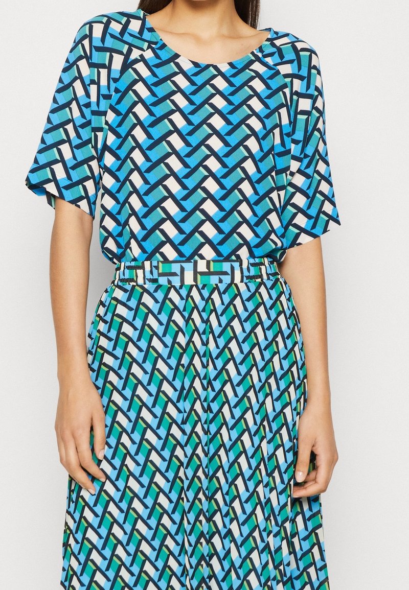 Femme portant un haut à manches courtes et un pantalon assortis avec un motif géométrique de maillons en bleu, vert, noir et blanc.