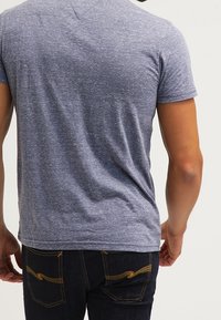 Grå kortärmad t-shirt med en strukturerad tygkvalitet; kombinerad med mörka jeans med gula sömnadsdetaljer. Bakifrån visas en figurnära silhuett.