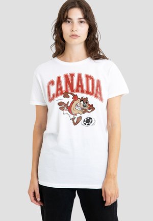 Junge Frau trägt ein weißes T-Shirt mit großem roten "CANADA"-Text und einer Cartoonfigur, die einen Fußball auf einem schlichten grauen Hintergrund kickt.