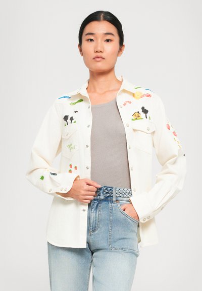Jeune femme portant une chemise blanche à boutons avec des motifs brodés colorés, un débardeur gris et un jean taille haute bleu clair avec une ceinture tressée.