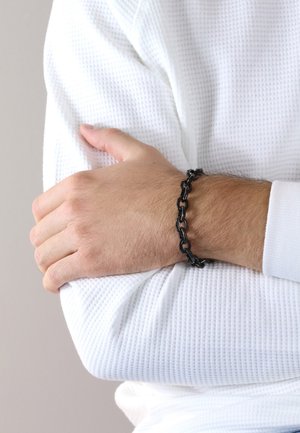 Black metal chain bracelet on a wrist, featuring linked segments. The wrist is covered with a textured white long-sleeve shirt.
