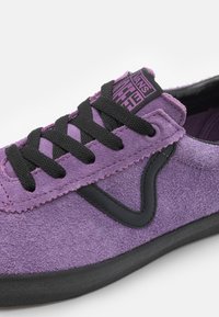 Ténis em suede roxo com detalhes em preto, apresentando atacadores pretos e uma etiqueta com o logo. Superfície suave e formato arredondado com sola de borracha.