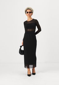 Juodas ilgos rankovės suknelė su permatomu pamušalu, prigludusia suknelės dalimi ir tiesiu siluetu, derinama su juodais aukštakulniais ir maža rankine.