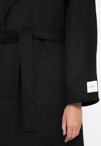 Black wool blend robe with a tie waist, large sleeves, and a white brand label on the sleeve. Soft texture and minimalist design.