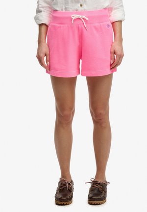 Donna indossa pantaloncini rosa acceso con coulisse, camicia bianca a maniche lunghe arrotolate e scarpe marroni con lacci, in piedi contro uno sfondo bianco.