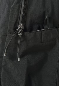 Musta kangasvetoketjun yksityiskohta, jossa on ristikkäiset metallivetimet, lisäremmejä ja neulatulla taskukuvioinnilla. Näkyvä teksturoitu pinta.