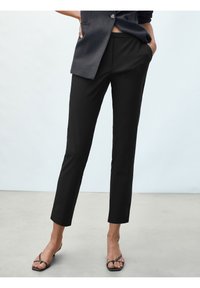 Kvinna klädd i svarta skräddarsydda ankellånga byxor, mörkgrå ärmlös väst och svarta remsandaler med klack, stående med händerna i fickorna.