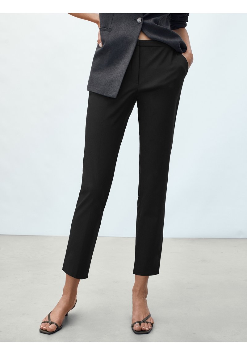 Kvinna klädd i svarta skräddarsydda ankellånga byxor, mörkgrå ärmlös väst och svarta remsandaler med klack, stående med händerna i fickorna.