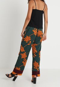 Black tank top with thin straps, paired with loose green pants featuring orange floral pattern and polka dots; maroon trim at cuffs.