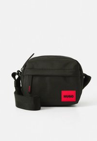 Melna neilona somiņa pāri plecam ar regulējamu siksnu, rāvējslēdzēja aizdari un sarkanu taisnstūra formas logotipa uzšuvumu priekšpusē. Kompakts dizains ar teksturētu audumu.