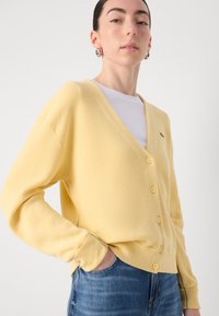 Žlutý kardigan s výstřihem do V, pěti knoflíky, ribbovanou texturou a zelenými akcenty. V kombinaci s bílou košilí a džíny.