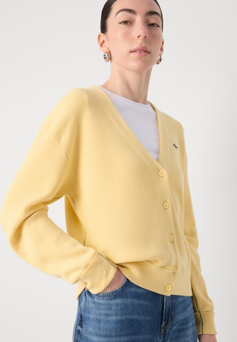 Gelbes Cardigan mit V-Ausschnitt, fünf Knöpfen, gerippter Textur und grünen Akzenten. Kombiniert mit einem weißen Hemd und Jeans.