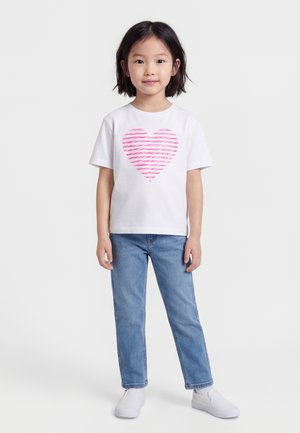 Ragazza giovane con capelli neri che indossa una t-shirt bianca con un cuore a righe rosa, jeans blu e scarpe slip-on bianche, in piedi contro uno sfondo bianco.