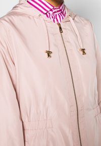 Ljust rosa huvjacka i slätt, glansigt material med dragkedja framtill och elastisk midja. Utrustad med detaljer i guldton och dragsnören.