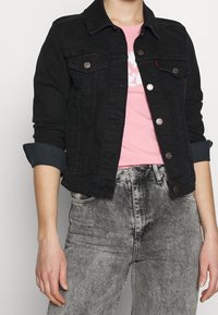 Černá džínová bunda s předními kapsami, kovovými knoflíky a ohrnutými rukávy, nošená přes růžové tričko s grafikou a šedé rifle s vysokým pasem.