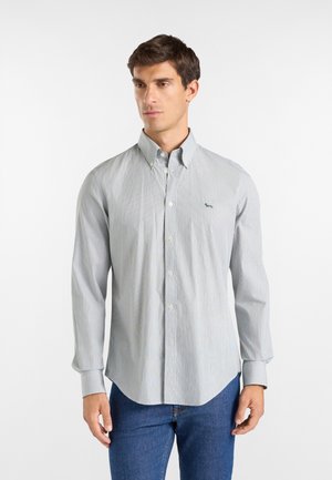 Hombre que lleva una camisa gris claro abotonada con un pequeño logo de perro en el pecho y vaqueros azules, de pie frente a un fondo blanco.