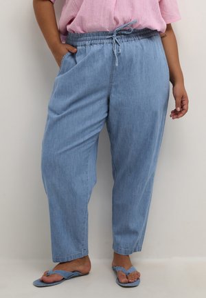 Person wearing light blue drawstring pants, pink top, and blue flip-flops, standing with one hand in pocket against plain wall.