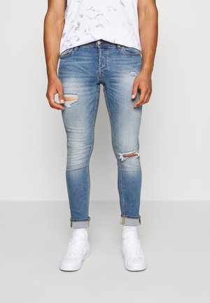 Verblasste blaue Denim-Jeans im Slim-Fit, mit Rissen an den Knien und einem rohen Saum. Kombiniert mit weißen Sneakers und Socken.