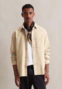 Beige textuur shirt met knoopsluiting, enkele borstzak, gedragen over een witte T-shirt, in combinatie met zwarte jeans en een gebreide sjaal.