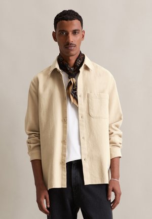 Beige textured shirt with button closure, single chest pocket, worn over a white t-shirt, paired with black jeans and a patterned scarf.